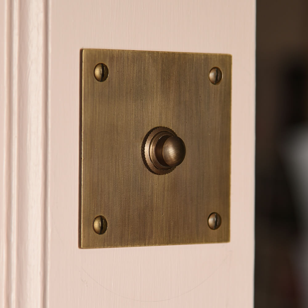 Distressed darkened brass square shaped door bell on a pink door frame