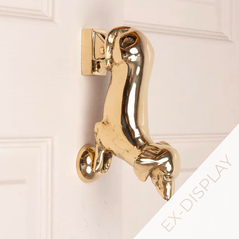 Solid brass dachshund door knocker with the front feet acting as a knocker on a pale pink door with a watermark and ex display text in the corner 
